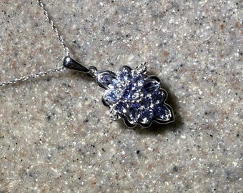 May include: A silver-toned necklace featuring a pendant shaped like a cluster of leaves or petals. The pendant is set with multiple small, light blue, oval-cut stones and tiny clear accent stones, suspended from a delicate, thin silver-toned chain.