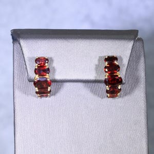 May include: A pair of gold hoop earrings, each featuring a vertical row of four oval-cut deep red garnet gemstones. The stones are prong-set in a polished gold metal frame, creating a continuous line of faceted red gems on each earring.