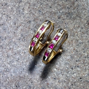 May include: A pair of gold hoop earrings featuring a channel setting with alternating round-cut pink and clear stones. The earrings have a hinged closure mechanism and a polished metallic finish, showcasing a symmetrical design with three pink stones.