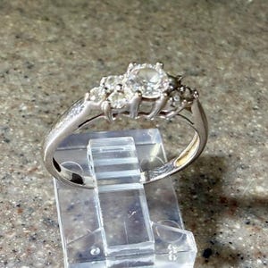 May include: A silver ring with a large, round, clear stone in the center, flanked by smaller stones. The ring is set on a clear plastic display stand, showcasing its design and sparkle. The ring's band is also silver.
