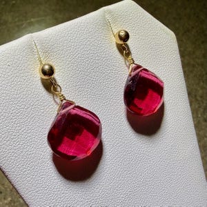 May include: A pair of gold-toned drop earrings. Each earring features a small gold ball at the top, connected to a faceted, teardrop-shaped, deep red gemstone. The gemstones are suspended by delicate gold wire.