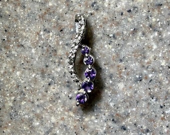 May include: A silver pendant with a curved design. The pendant features a line of small, clear stones on one side and a series of five round, deep purple gemstones on the other. The pendant is set against a speckled gray background.