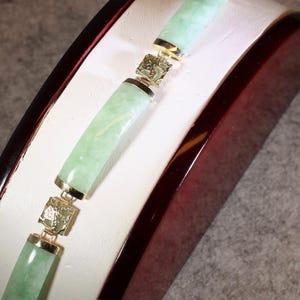 May include: A bracelet featuring alternating cylindrical light green jade-like stone segments and square gold-toned metal links. The metal links are textured with intricate engraved patterns, connecting the smooth, polished stone pieces together.