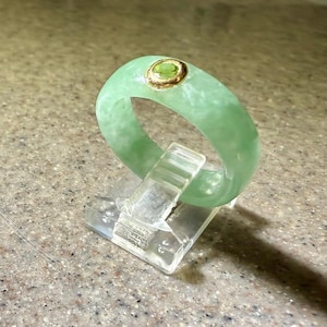 May include: A pale green jade band ring featuring a single round, faceted gemstone set in a polished gold-tone bezel. The ring has a smooth, rounded profile and a mottled, semi-translucent appearance, displayed upright on a clear plastic stand.