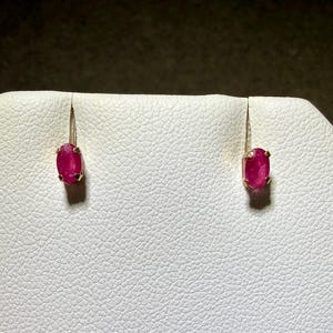 May include: A pair of earrings featuring oval-shaped, deep pinkish-red rubies set in gold metal. Each gemstone is secured by four small prongs, showcasing a faceted surface that reflects light. The earrings are displayed vertically on a textured surface.