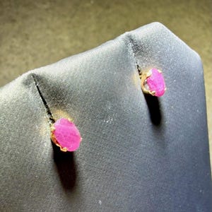 May include: A pair of stud earrings featuring oval-shaped, vibrant pink gemstones set in gold-toned metal prongs. The stones have a textured, natural surface appearance and are displayed on a dark, angled stand to highlight their color.
