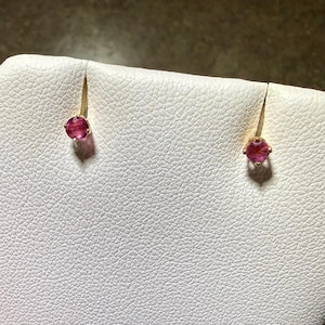 May include: A pair of stud earrings featuring round, faceted, deep pink gemstones set in gold-toned metal prongs. Each earring is secured to a white, textured display surface, showcasing the vibrant color and circular cut of the stones.