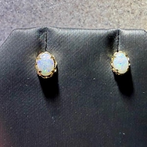 May include: A pair of oval-shaped stud earrings featuring iridescent white opals set in yellow gold prongs. Each opal displays flashes of blue and green color play within the stone, secured by a simple metal setting that holds the gem firmly.