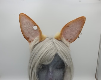 May include: A pair of cosplay fox ears with a light brown and tan color scheme. The ears are adorned with small silver charms, including a paw print and a heart. The ears are attached to a headband.