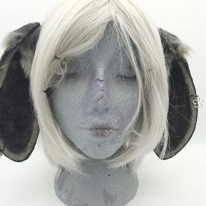 May include: A pair of dark gray, fuzzy animal ears attached to a headband, worn on a gray mannequin head with a short, straight white wig. The ears feature a soft, textured surface and are adorned with small silver-toned charms, including a flower shape.