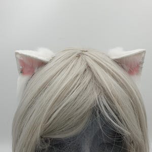 May include: A pair of white faux fur cat ears attached to a headband, worn over straight platinum blonde hair. The ears feature soft pink inner coloring, with a small silver stud earring on the left side and two silver hoop rings on the right.