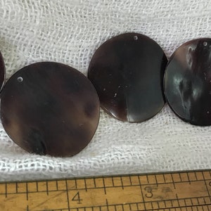 May include: Five round, dark purple, iridescent, shell-like discs with a small hole in the center. The discs are arranged in a row on a white fabric background.