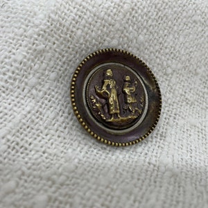 May include: A round, gold-colored button with a raised design of two figures in a scene. The button has a textured, beaded edge.
