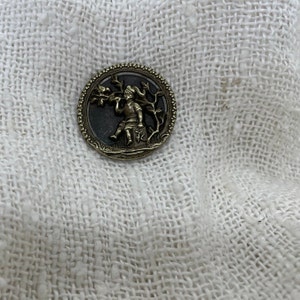 May include: A round, silver-toned button with a black background. The button features a raised design of a child holding a branch with leaves. The button has a decorative border.
