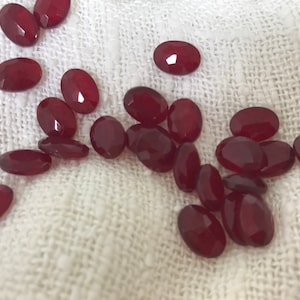 Pode incluir: Uma coleção de pedras preciosas vermelhas de forma oval. As pedras preciosas estão espalhadas sobre uma superfície de tecido branco.
