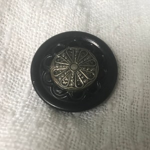 Pode incluir: Um botão preto com um centro de filigrana prateada. O botão tem uma borda ondulada e um design floral decorativo. O botão está sobre um fundo branco texturizado.