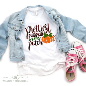 May include: White t-shirt with the text "Prettiest Pumpkin in the Patch" and a pumpkin graphic. The shirt is paired with a light blue denim jacket and pink sneakers with white laces.