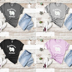 May include: Four different color t-shirts with a white graphic of a Pomeranian dog with the text "Pomeranian mama" on the front. The shirts are displayed on a light wood background with a pair of jeans and a pair of white sneakers next to each shirt.