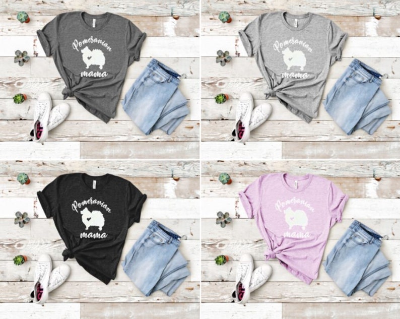 May include: Four different color t-shirts with a white graphic of a Pomeranian dog with the text "Pomeranian mama" on the front. The shirts are displayed on a light wood background with a pair of jeans and a pair of white sneakers next to each shirt.