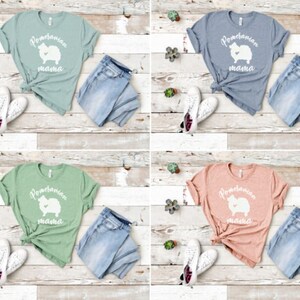 May include: Four different color t-shirts with a white Pomeranian silhouette and the text "Pomeranian mama" on the front. The shirts are displayed on a light wood background with blue jeans and white sneakers.