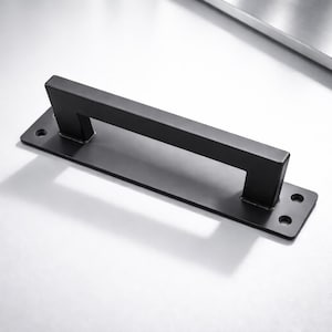 May include: A black, rectangular door pull with a square handle. The pull has a flat base with pre-drilled holes for mounting. The handle is a simple, modern design, and the hardware is made of metal.