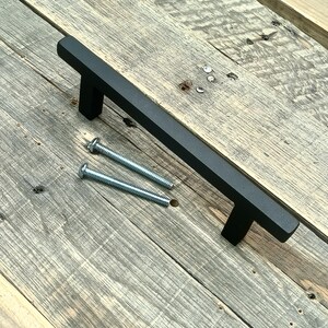 May include: A black cabinet pull with a rectangular bar and two screws, set against a weathered wooden surface. The hardware is a matte black finish, designed for furniture or cabinetry. The screws are silver.