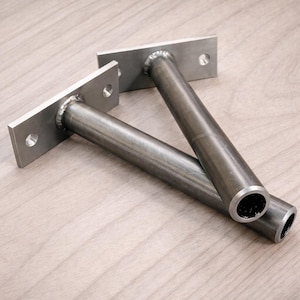 May include: Two metal shelf brackets. Each bracket has a rectangular mounting plate with two holes and a cylindrical support rod. The brackets are made of silver-coloured metal. Designed for supporting shelves, these brackets are suitable for various home decor projects.