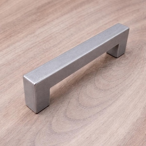 May include: A silver-colored cabinet pull with a rectangular bar and two square-shaped feet. The hardware is made of metal and is designed for easy installation on drawers or cabinet doors. The pull is shown on a light brown wooden surface.