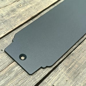 May include: A dark gray metal tag with a decorative edge and a hole for hanging. The tag has a textured surface and is rectangular with a rounded corner. The tag is shown on a wooden surface.
