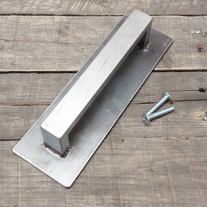 May include: A silver metal door handle with a rectangular plate and a raised bar. The handle is shown on a wooden surface with two screws.