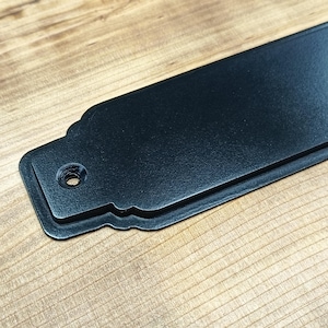 May include: A close-up of a black metal sign with a textured surface. The sign is rectangular with a decorative edge and a single hole for mounting. The sign is set against a wooden background.