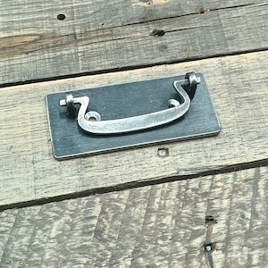 May include: A rectangular, dark gray metal drawer pull with a curved, silver-colored handle. The handle is attached to the rectangular base with two curved supports. The hardware is designed for furniture and cabinetry.