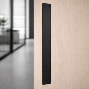May include: A black, rectangular door pull mounted vertically on a light wood door. The pull is made of a solid, matte black material and is secured with screws at the top and bottom.