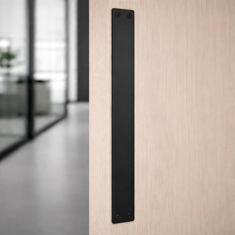 May include: A black, rectangular door pull mounted vertically on a light wood door. The pull is made of a solid, matte black material and is secured with screws at the top and bottom.
