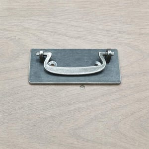 May include: A rectangular, dark gray metal drawer pull with a curved, silver handle. The handle is attached to the rectangular base with two decorative, curved brackets. The hardware is designed for furniture and cabinetry.