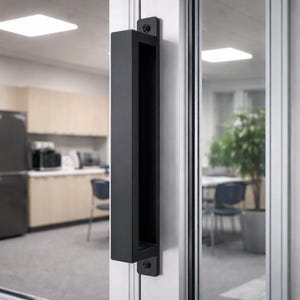 May include: A black rectangular door pull handle mounted on a white door frame. The handle is a long, hollow rectangle with a smooth, matte finish. The handle is attached to the door with two small, square black plates and screws.