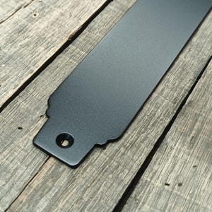 May include: A close-up of a black metal strap with a decorative edge and a circular hole. The strap is set against a weathered wooden surface, showcasing its textured finish and design.