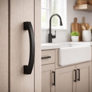 May include: A black, curved cabinet pull handle mounted on a light wood cabinet door. The handle has a modern, minimalist design. The background shows a kitchen with a white sink and black tap.