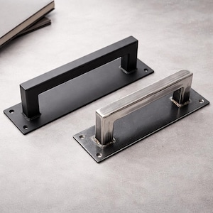 May include: Two metal door pulls. One is black, the other is silver. Each has a rectangular base with a square handle. The door pulls are shown on a gray surface. The handles are approximately 6 inches long.