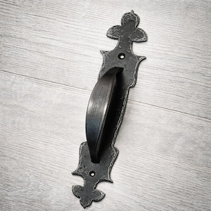 May include: A black, ornate door pull with a curved handle and decorative ends. The metal hardware has a distressed finish, and is designed for a rustic or vintage aesthetic. The door pull is mounted on a light gray wooden surface.