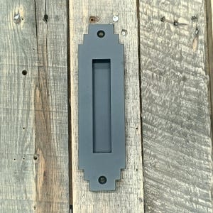 以下が含まれることがあります： 幾何学模様のデザインが施された、長方形のグレーの金属製埋め込みプル。風化した木製の板に取り付けられています。プルには細長い開口部があり、2本の黒いネジで固定されています。