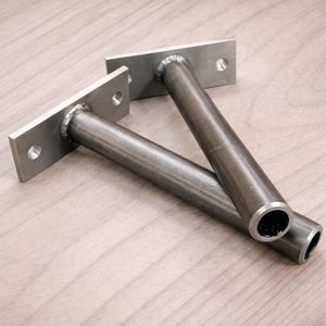 May include: Two metal shelf brackets with a rectangular mounting plate and a cylindrical support rod. Each bracket has two pre-drilled holes for mounting. The brackets are made of a silver-coloured metal and are designed for wall mounting.