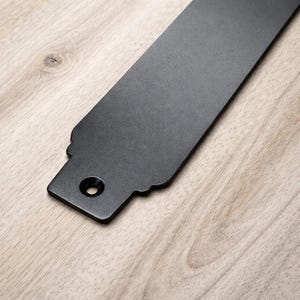 May include: A matte black metal object with a rectangular shape and a decorative edge. The object has a hole at one end and is set against a light wood grain surface.