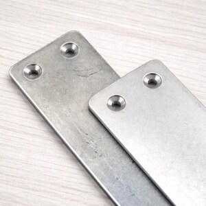 May include: Two rectangular metal plates with rounded corners and two pre-drilled holes each. The plates are a brushed silver color and appear to be made of stainless steel. One plate is larger than the other, and they are slightly overlapping.