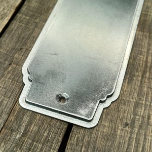 May include: A rectangular, galvanized metal plate with a decorative scalloped edge. The plate has a single hole near the bottom and sits on a weathered wooden surface. The metal has a shiny, reflective surface.