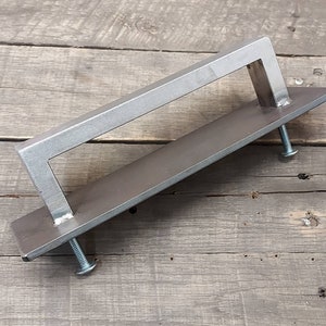 May include: A silver metal door handle with a rectangular shape and a raised bar. The handle is mounted on a flat metal plate.