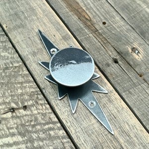 Peut inclure: Un bouton de meuble en métal argenté en forme d'éclat d'étoile avec un centre circulaire. Le bouton a une finition vieillie et est monté avec des vis. Le fond est une surface en bois.