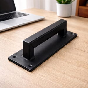 May include: A black metal door pull with a rectangular base and a rectangular handle. The door pull has a matte finish and is mounted on a wooden surface. The handle is approximately 6 inches long.