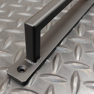 May include: Black metal door handle with a rectangular shape and a rounded edge. The handle is mounted on a flat metal plate with a screw visible.