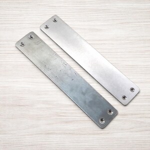 May include: Two rectangular metal plates with rounded corners and pre-drilled holes for screws. The plates are silver in color and have a brushed metal finish. They are lying on a light wood grain surface.